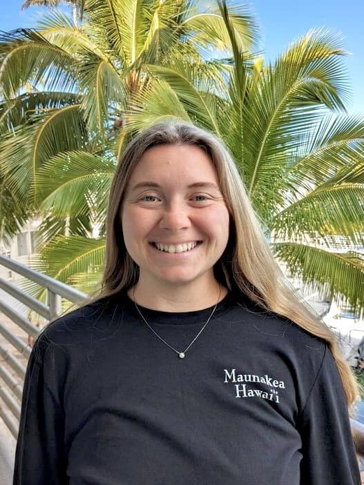 Smiling woman with long hair in a "Maunakea Hawai'i" shirt stands in front of palm trees outdoors.