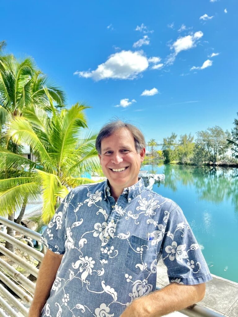 Smiling man in a floral shirt stands by a tropical waterfront with palm trees and blue sky in the background.