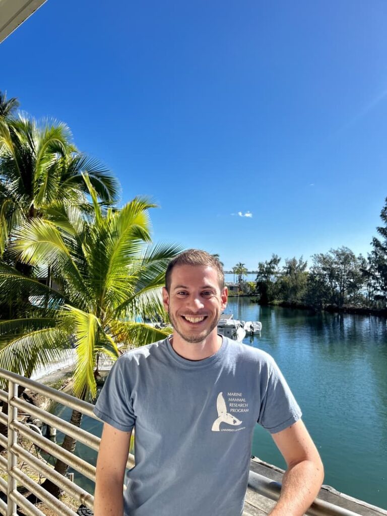 A man smiles on a balcony by palm trees and water under a clear blue sky on a sunny day.