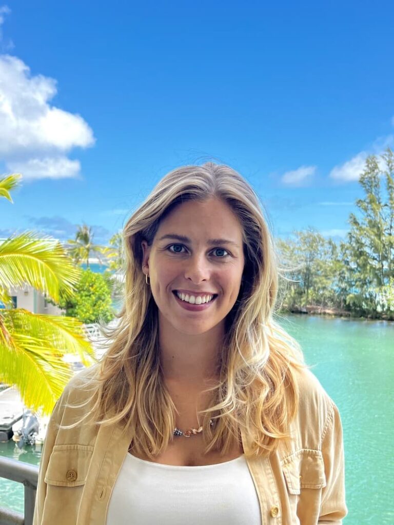 Smiling woman with blonde hair stands outside by turquoise water, palm trees, and a bright blue sky.