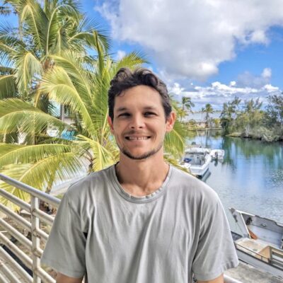 Smiling man in a gray shirt stands by a railing with palm trees and boats on the water in the background.