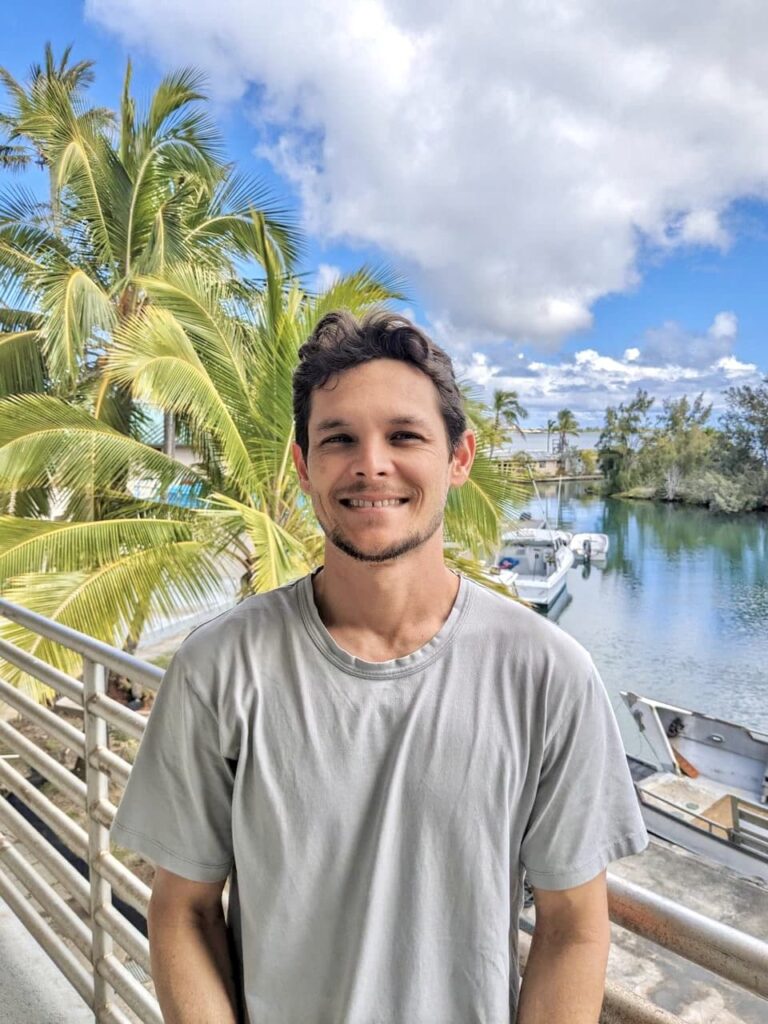 Smiling man in a gray shirt stands by a railing with palm trees and boats on the water in the background.