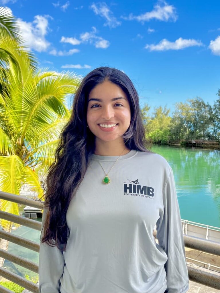 Smiling woman in a HIMB shirt stands by water with palm trees and blue sky in the background.
