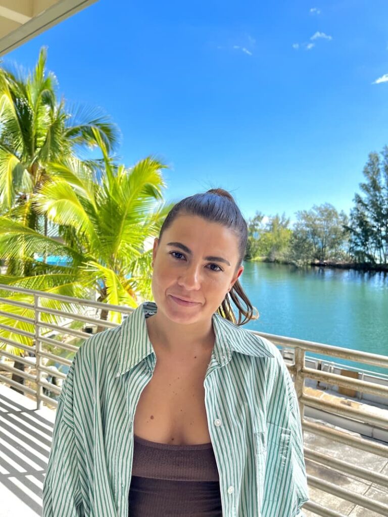 Woman in a striped shirt stands by a railing with water, palm trees, and blue sky in the background.