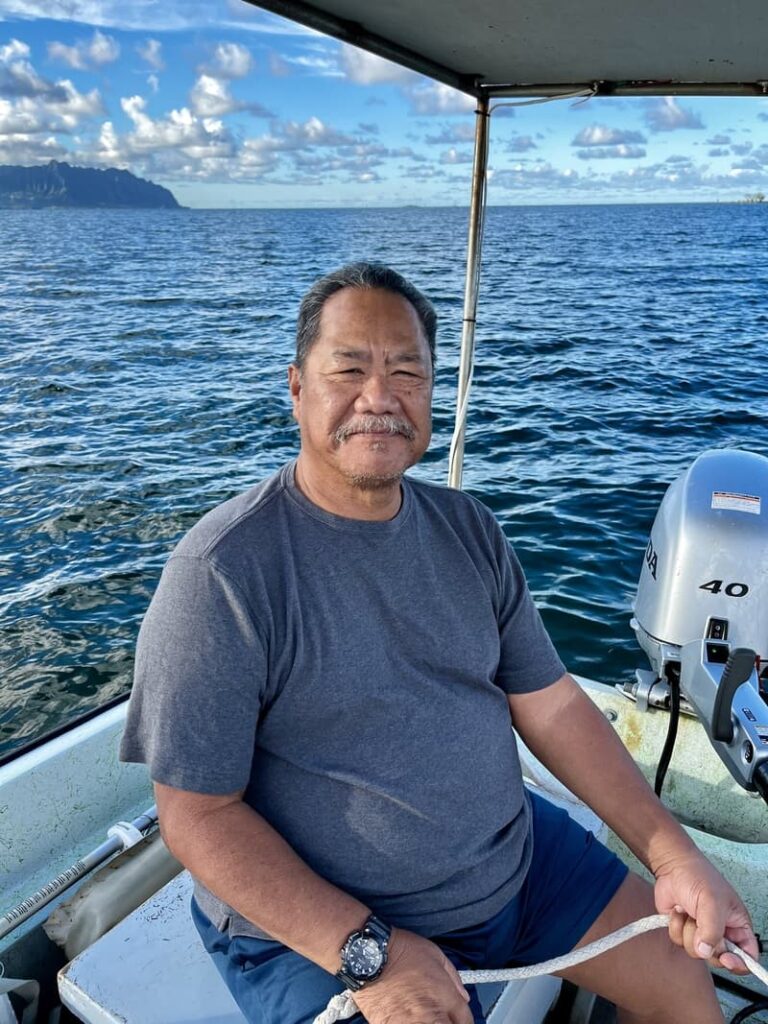 Man sitting on a boat holding a rope, with ocean and distant coast under a partly cloudy sky.