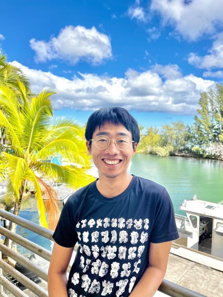 Smiling person in glasses stands by water with palm trees and boats under a bright blue sky with clouds.