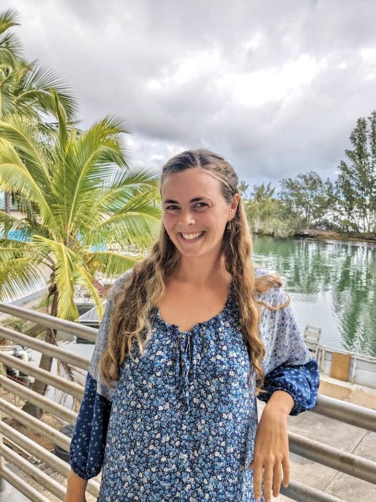 A woman in a blue floral dress smiles by a railing, with palm trees and water in the background.