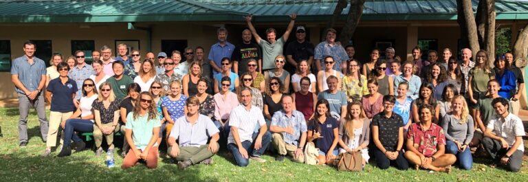 A large group of people pose and smile for a group photo outside on a sunny day, in front of a building.