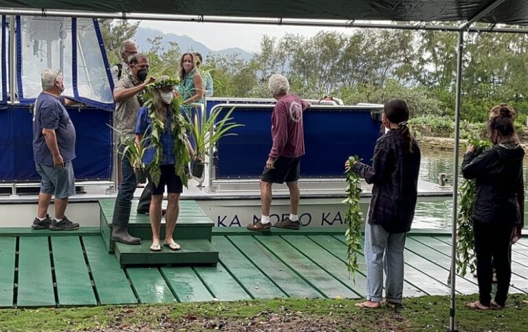 People participate in a traditional ceremony by a boat, some holding leafy garlands, under a canopy outdoors.