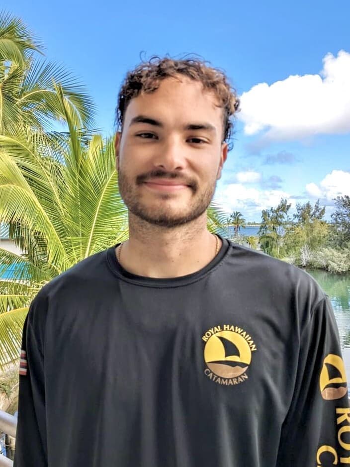 A young man in a black "Royal Hawaiian Catamaran" shirt stands outside with palm trees and blue sky behind him.