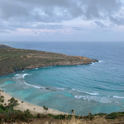 Scenic view of a crescent-shaped bay with clear blue water, sandy beach, and lush green hills under a cloudy sky.