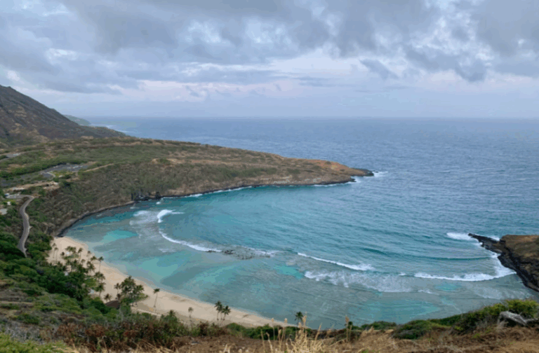 Scenic view of a crescent-shaped bay with clear blue water, sandy beach, and lush green hills under a cloudy sky.