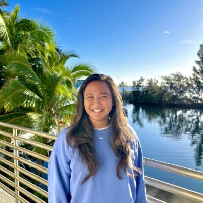 Woman smiling by a railing with palm trees and a calm lake under a clear blue sky in the background.