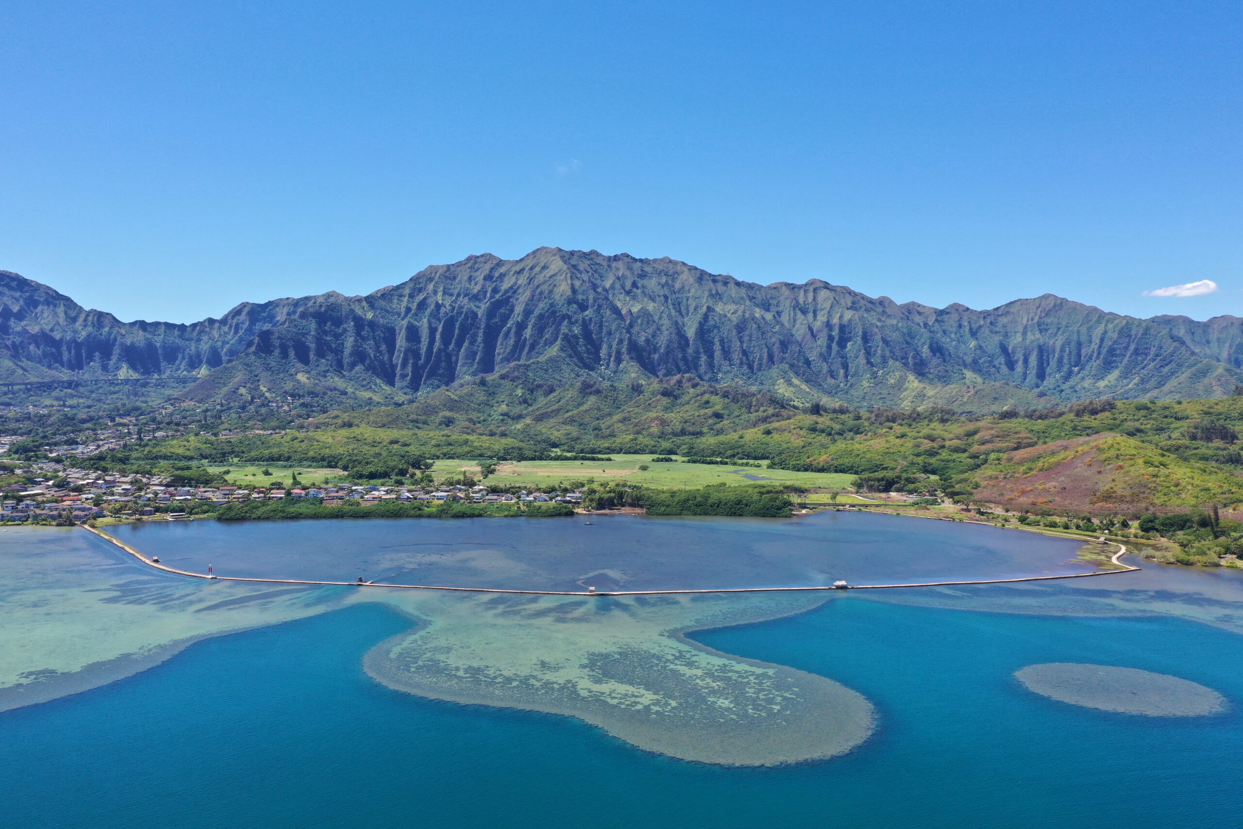 A scenic aerial view of a blue bay, sandbar, and lush green mountains under a clear sky.