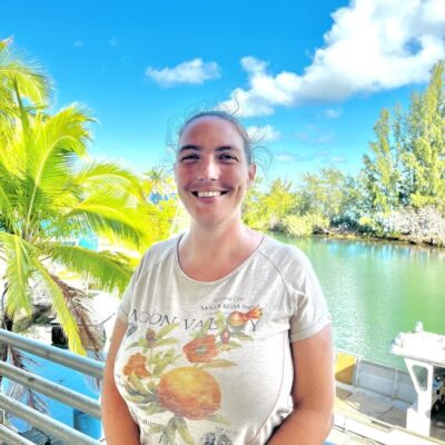 Smiling person stands by water, with palm trees and boats in the background under a bright blue sky.