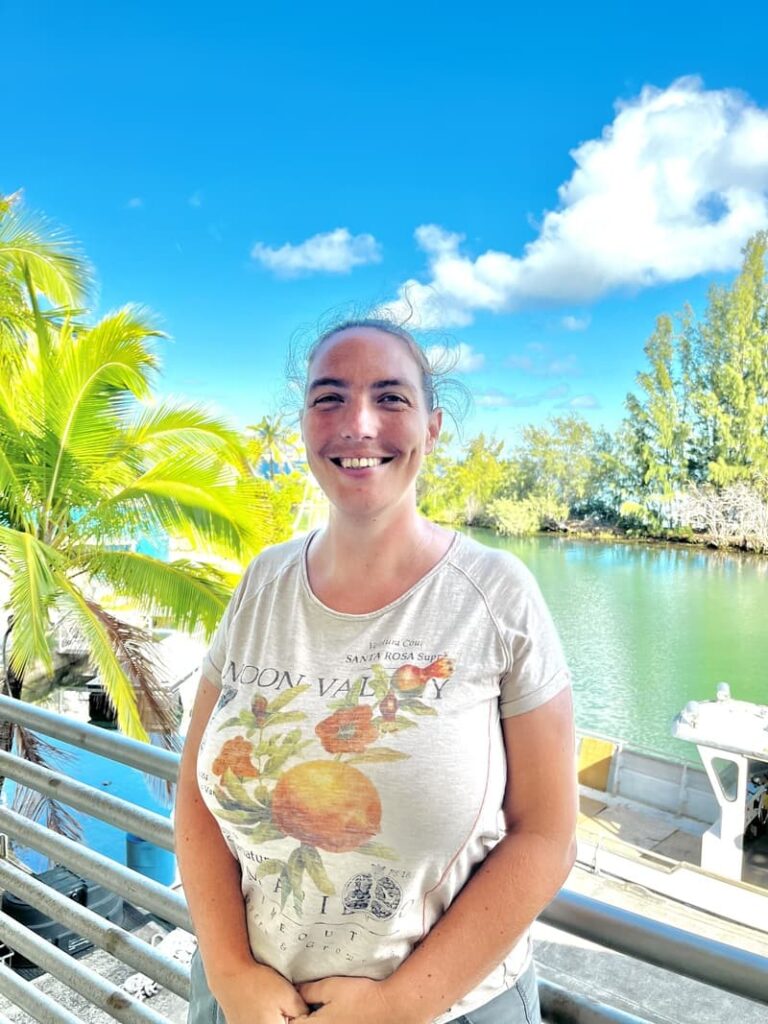 Smiling person stands by water, with palm trees and boats in the background under a bright blue sky.