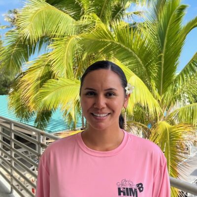 A woman in a pink shirt stands smiling in front of palm trees on a sunny day.