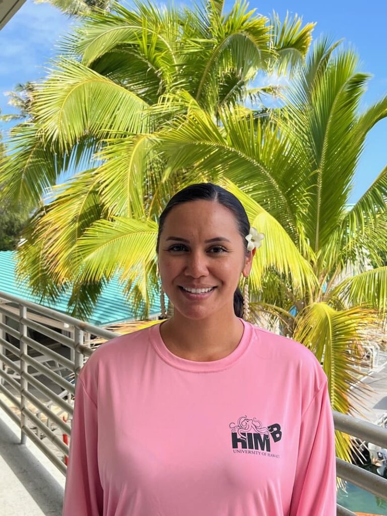 A woman in a pink shirt stands smiling in front of palm trees on a sunny day.