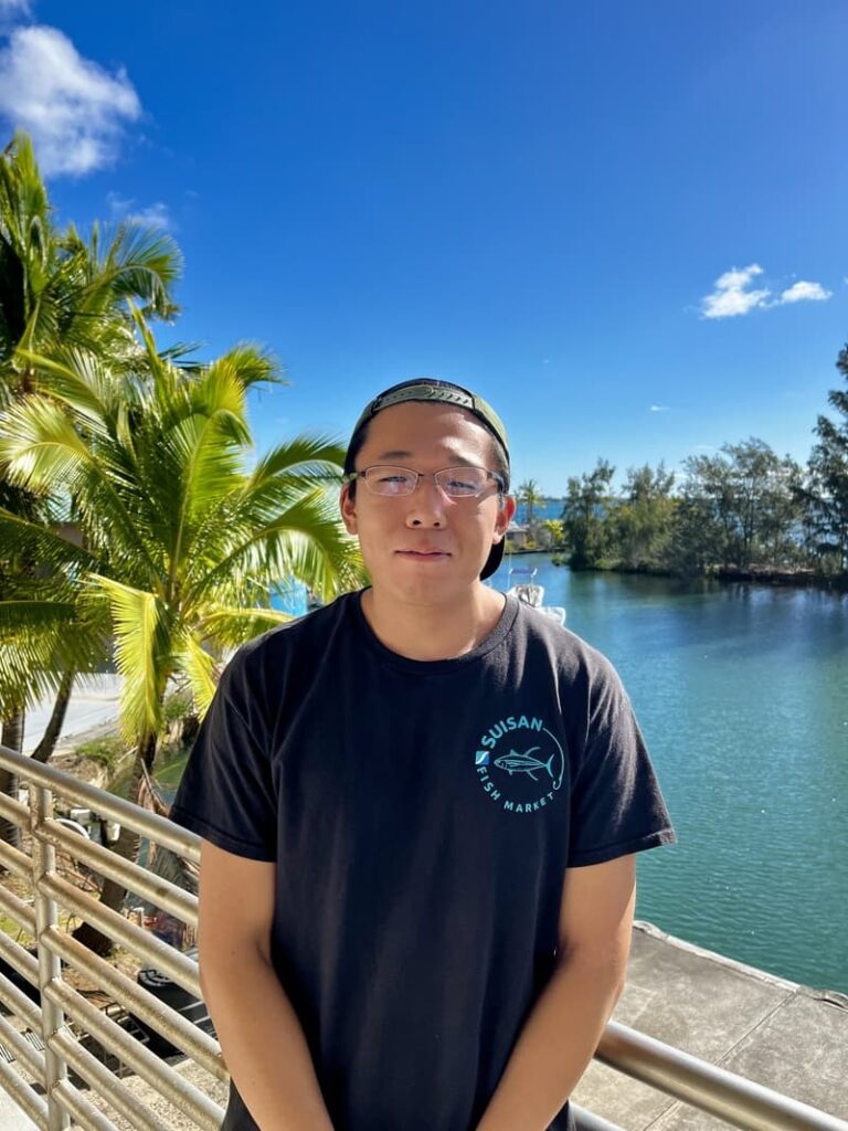 A person in glasses and a dark shirt stands by water and palm trees under a clear blue sky.