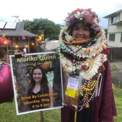 Young woman in graduation gown and lei stands beside a sign with her photo announcing a drive-by celebration.