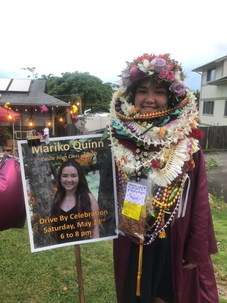 Young woman in graduation gown and lei stands beside a sign with her photo announcing a drive-by celebration.
