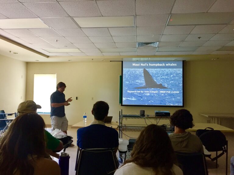 A person presents a slideshow on Maui Nui’s humpback whales to an audience in a classroom.