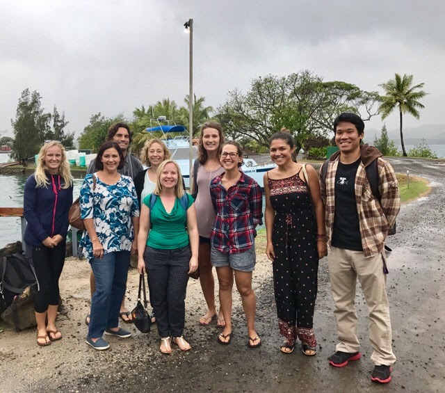 A group of nine people stand and smile together outside on a cloudy day near water and palm trees.