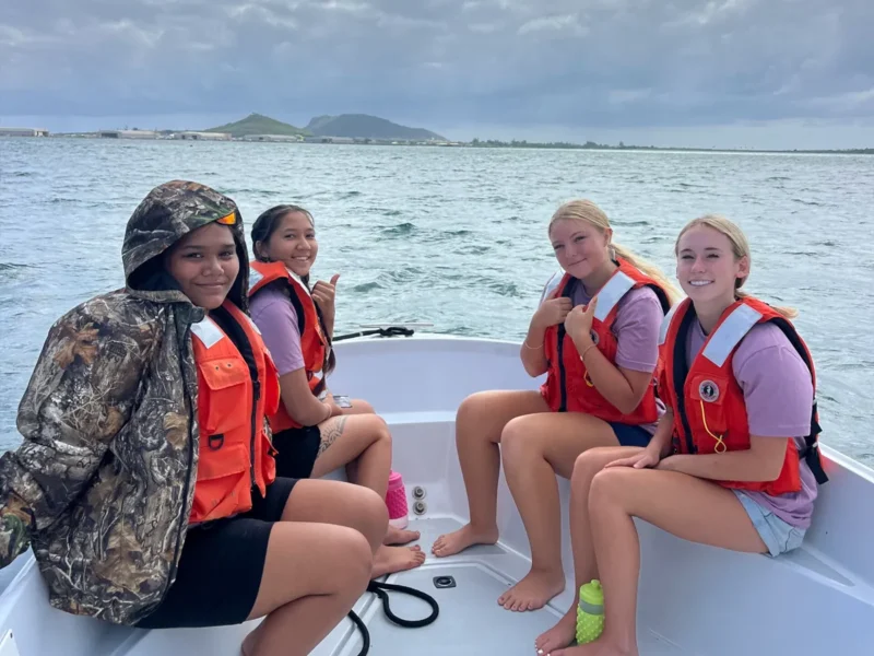 Four girls in life vests sit at the front of a boat on the water, smiling with an island in the background.