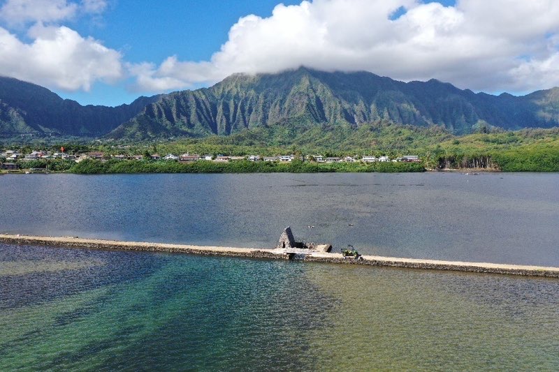 A stone walkway extends over calm water with green mountains and clouds in the background.