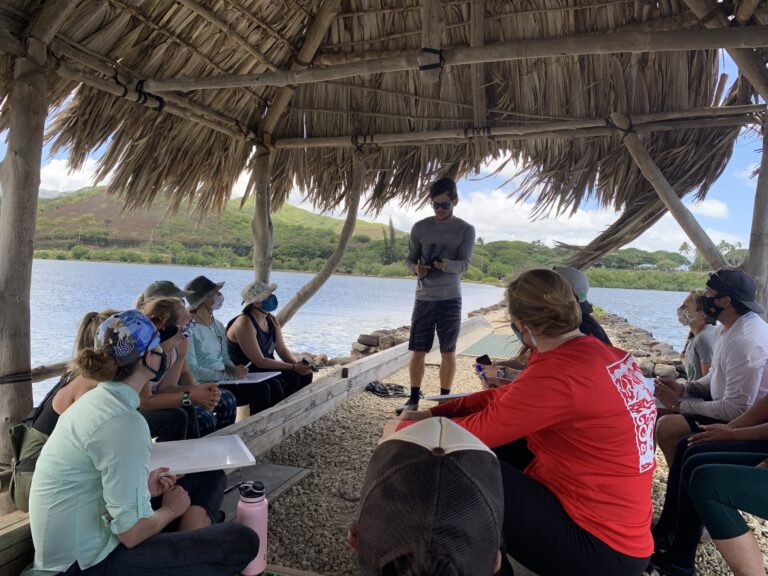 A group sits under a thatched shelter by the water, listening to a person standing and speaking outdoors.
