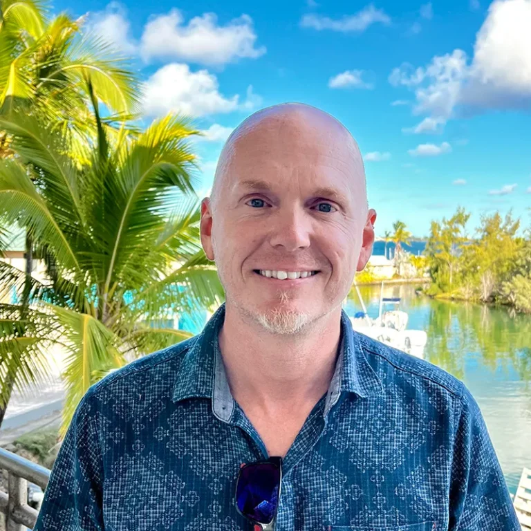 Smiling man stands outdoors by palm trees and water under a bright blue sky with scattered clouds.