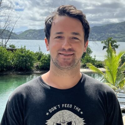 Man with dark hair and beard smiles outdoors by water, trees, and mountains, wearing a black "Don't Feed the" t-shirt.