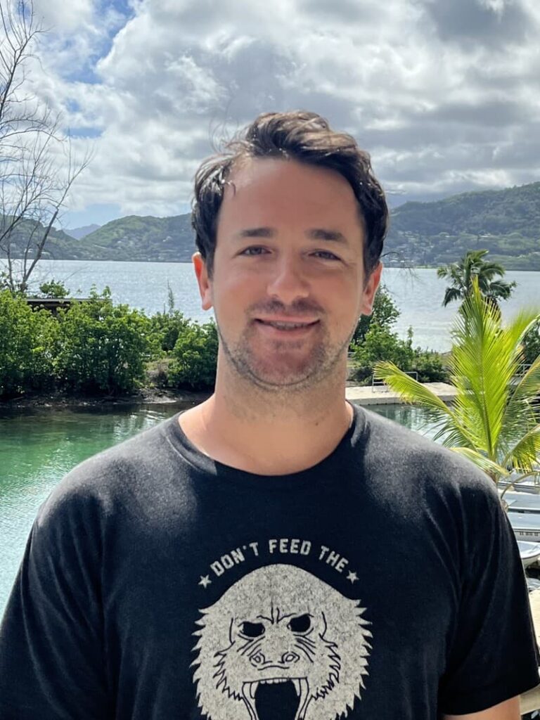 Man with dark hair and beard smiles outdoors by water, trees, and mountains, wearing a black "Don't Feed the" t-shirt.