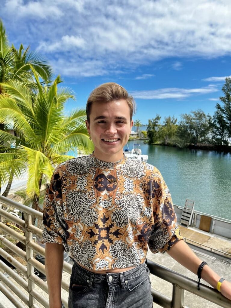 Smiling person in a patterned shirt stands by water, palm trees, and blue sky in the background.