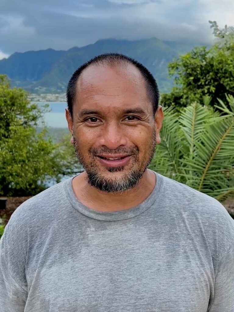 Smiling man in a gray shirt stands outdoors with mountains and greenery in the background.