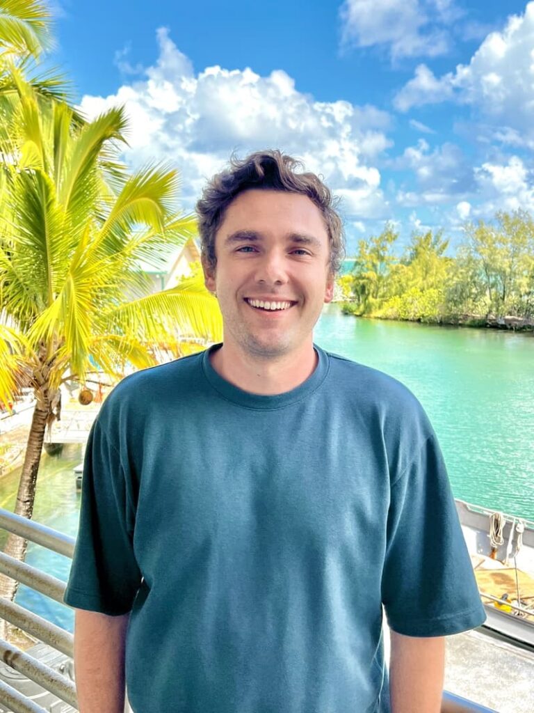 Smiling man in a blue shirt stands by a tropical river with palm trees and blue sky in the background.