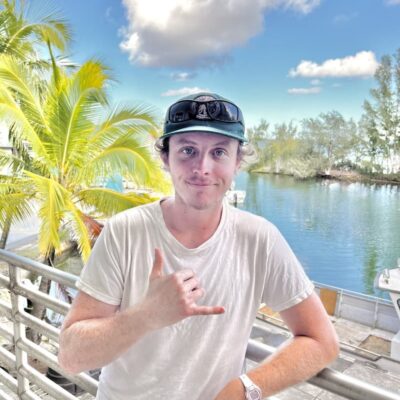Man in white shirt making shaka sign, standing by railing with palm trees and water in the background.