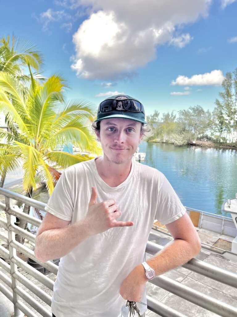 Man in white shirt making shaka sign, standing by railing with palm trees and water in the background.