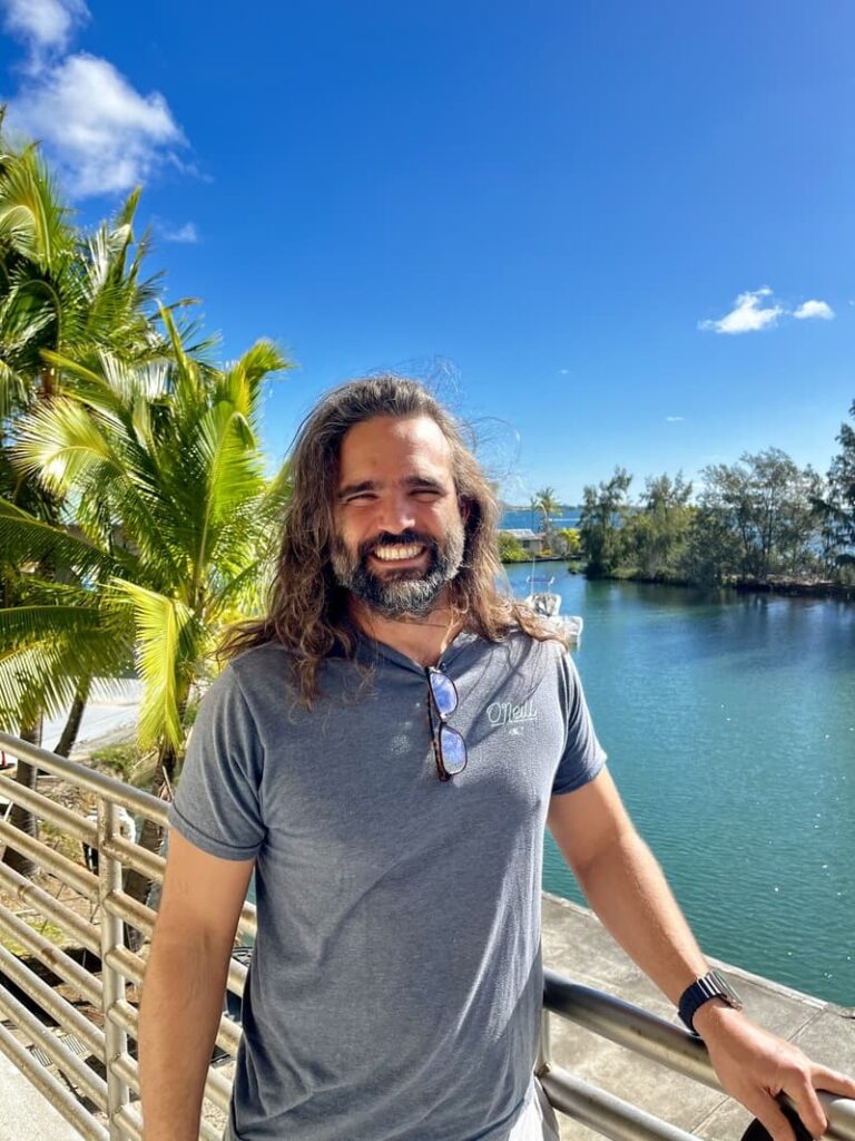 Man with long hair and beard smiling by a waterfront with palm trees under a bright blue sky.