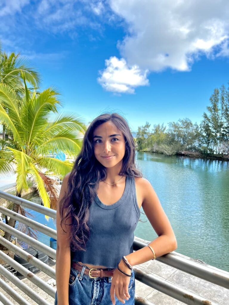 Woman with long hair in a tank top stands by a railing, with water, trees, and blue sky in the background.