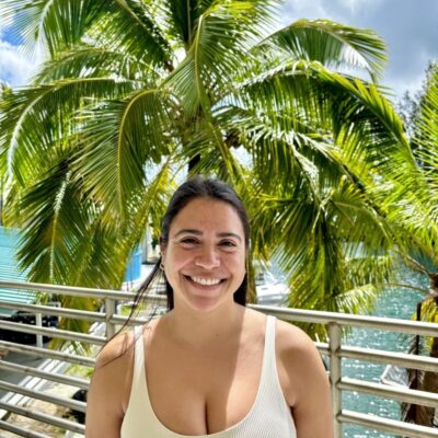 Woman smiling in front of palm trees and water on a sunny day, wearing a white tank top.