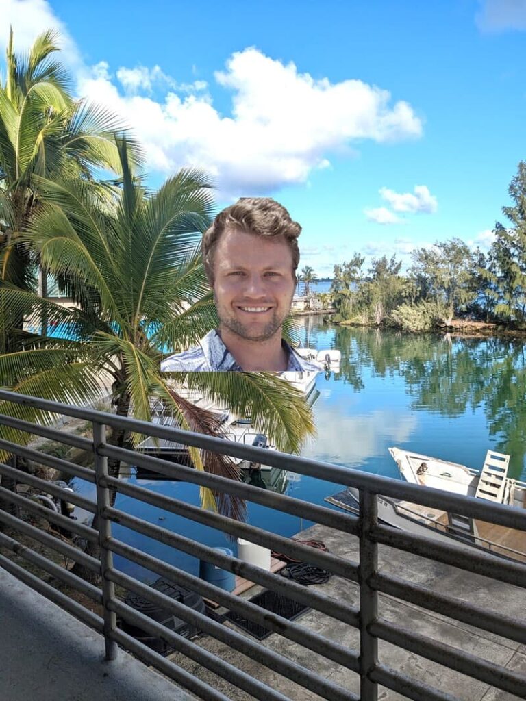 A man smiling near palm trees by a calm lake with boats, viewed from behind a metal railing.