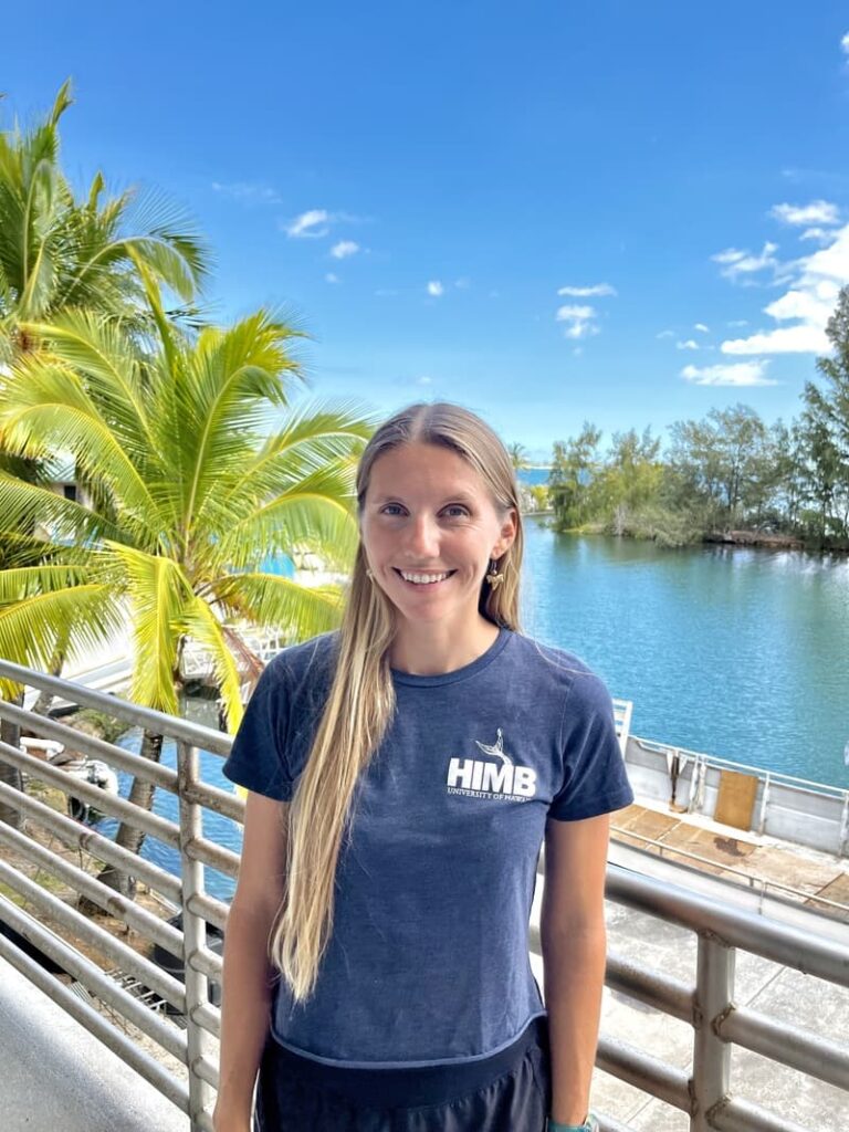 Smiling woman with long hair stands by water and palm trees, wearing a navy "HIMB" t-shirt on a sunny day.