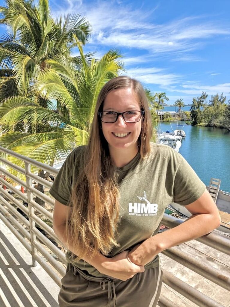 Smiling woman in glasses and HIME shirt stands by water with boats and palm trees on a sunny day.