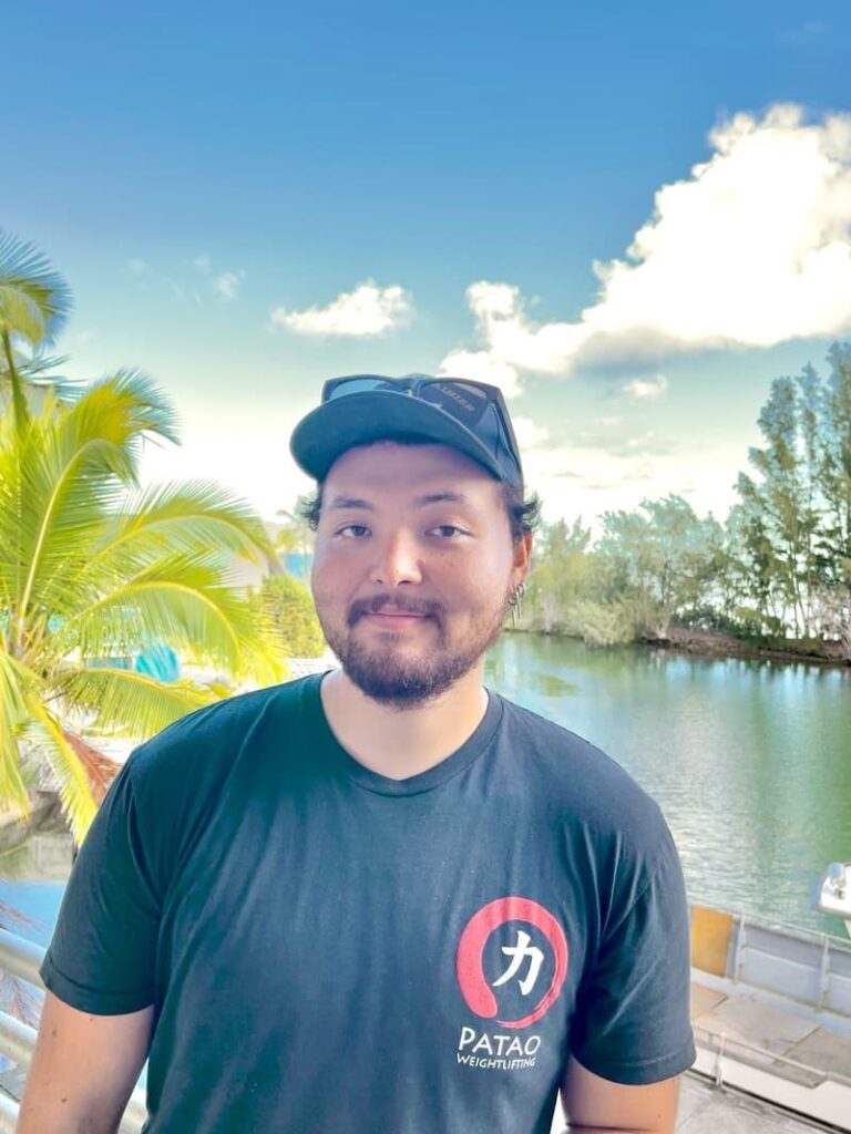 Man with a beard and cap stands by water and palm trees, wearing a black "PATAO" T-shirt, smiling at camera.