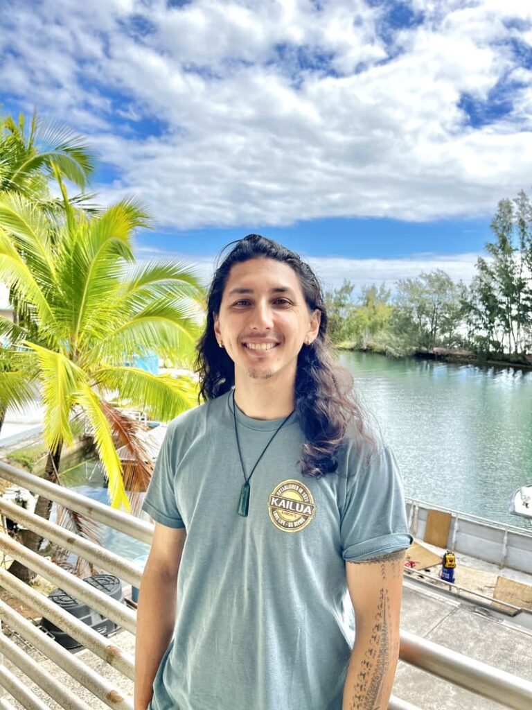 Smiling man with long hair stands by a riverside railing, palm trees and blue sky in the background.