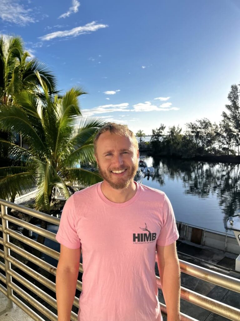 Smiling man in a pink Himb shirt stands by a railing with palm trees and water in the background.
