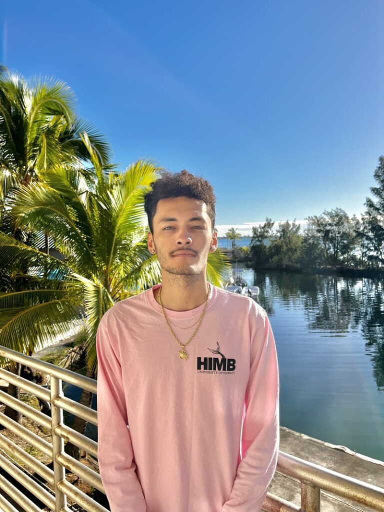 A young man in a pink shirt stands by water, with palm trees and a clear blue sky in the background.