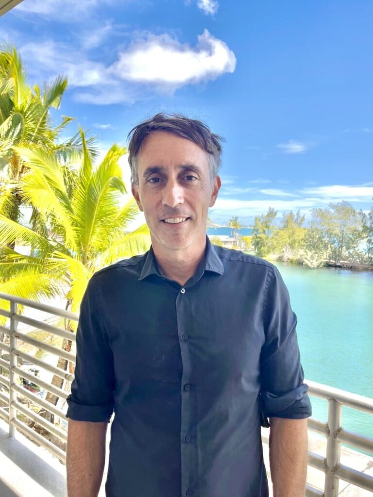 Man in a black shirt standing on a balcony with palm trees and blue water in the background.
