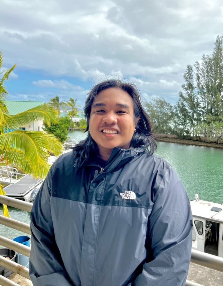 Person in a dark jacket smiling by a railing, with water, trees, and boats in the background.